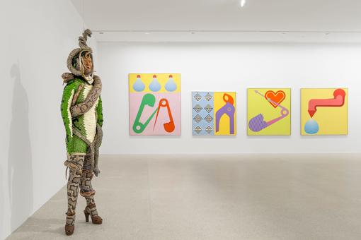 Exhibition view: in the foreground a figure in snakeskin and entwined with snakes, in the background four colorful, bright paintings with everyday objects, e.g. pliers, light bulbs