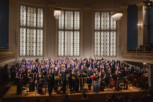 The photo shows the Vienna Bach Soloists and the BAchCHorWien on the stage of the Mozart Hall in the Vienna Konzerthaus, with colorful Art Nouveau stained glass windows behind them