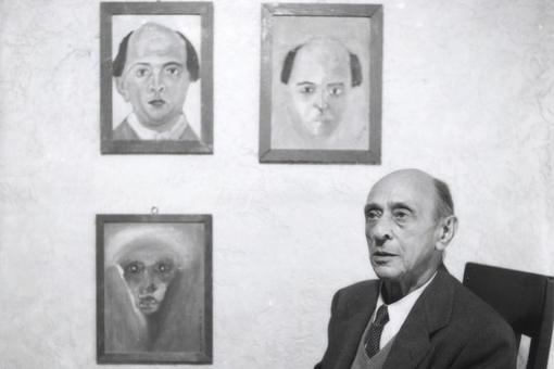 The black and white photo shows the composer Arnold Schönberg seated, in the background on the wall are three pictures painted by him, two of which are self-portraits