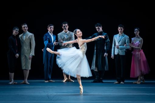 A ballerina in a white tulle dress on lace in the foreground, dancers in modern costumes / suits in the background