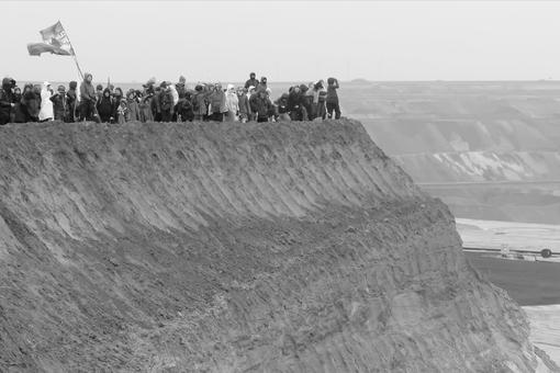 The black and white photo shows a protest rally of people at a steep precipice in the open-cast coal mining area of Lützerath
