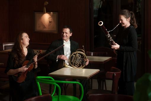 Foto von drei Mitgliedern der Wiener Symphonikern in einem Wiener Beisl: ein Herr in Frack mit einem Horn in Händen, eine Geigerin mit Violine und eine Frau mit einem Fagott in Händen