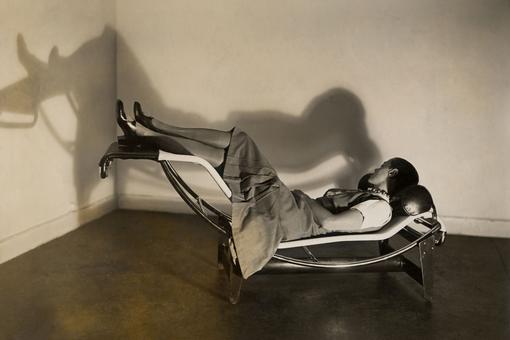 Black and white photo of the designer Charlotte Perriand lying on a chaise longue, a design piece made of metal with upholstery.
