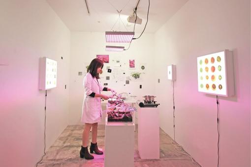 The photo shows a woman in a white work coat in a larbor-like situation with plants, illuminated screens on the walls, pink Led lighting, laboratory equipment