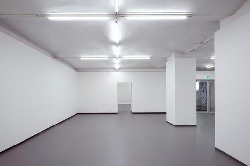 The photo shows an empty exhibition room with pure white walls and neon lighting, in the background a glass entrance door and a passageway into another room