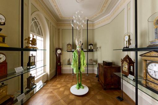 On display is a Biedermeier room with various historical clocks, in the center is a model in lime green by fashion designer Florentina Leitner