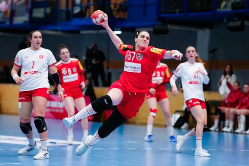 The photo shows a scene from a women's handball match, in the foreground a player in a red jersey taking a shot with the ball in her hand