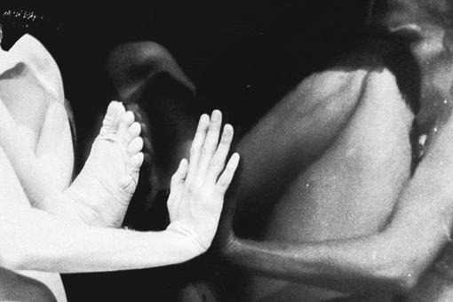 The black and white photo shows a scene as if underwater: a naked person and a person in a white jersey huddled together and one hand and one foot of each person meet through a pane of glass