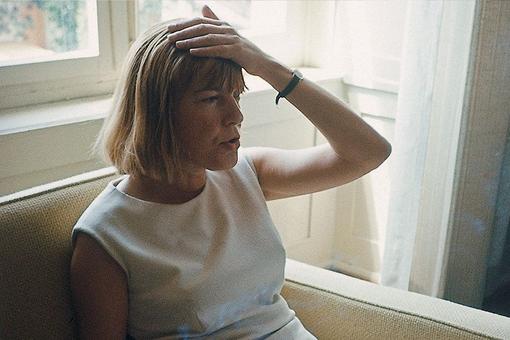 Photo of Ingeborg Bachmann sitting on a couch in a white dress and grabbing her head with her left hand