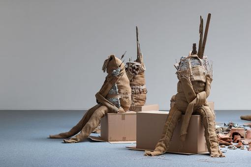 Photo of an installation consisting of three headless figures made of brown jute sitting on boxes made of brown cardboard