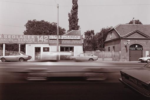 The black-and-white photo shows part of the busy Triester Strasse in Vienna 1982-83, with a car dealer's building and a tobacconist's in the background and moving cars in the foreground