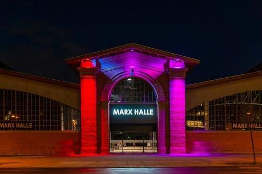 Photo of the main entrance of Marx Hall at night, bathed in red-pink light. The portal has a central round gate at the top, with two columns on the sides, and a gable roof above. Above the entrance the inscription Marx Hall