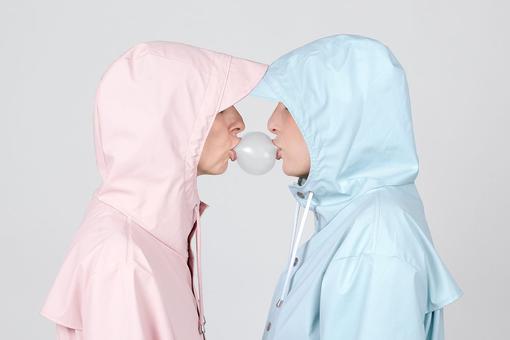The photo shows two people wearing a pink and a light blue hooded raincoat and standing opposite each other. There is a bubble of chewing gum between their kissing mouths
