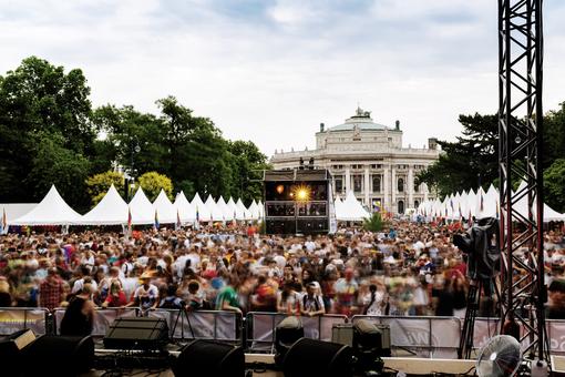 The photo shows Vienna's Rathausplatz, looking towards the Burgtheater from a stage, a crowd of people in front of the stage, white tents with pointed roofs at the edge