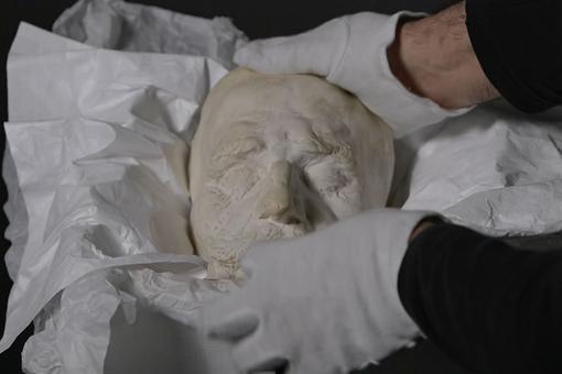 The photo shows hands in white gloves wrapping a light-colored death mask in white paper