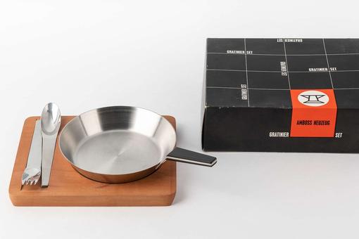 Stainless steel gratin set consisting of a small pan, a fork and a spoon on a wooden board with black cardboard packaging