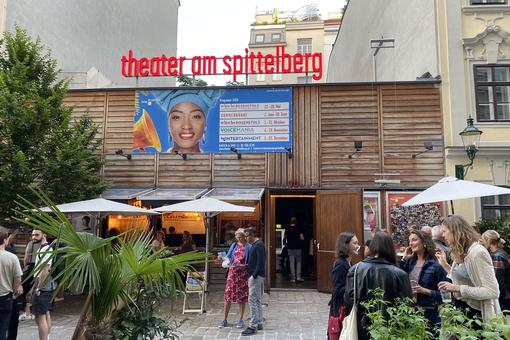 Exterior view of the Theater am Spittelberg, a wooden construction, in front of it audience in the break, deck chairs, umbrellas and plants