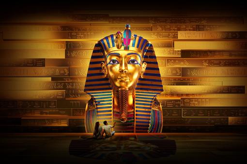 Image of the famous death mask of Tutankhamun in oversized representation, in the background a wall with hyroglyphs, in front of the mask sit a woman and a man