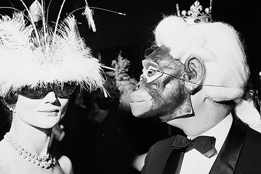 Black and white photo of elegant lady in feathered hat and black sunglasses accompanied by man in black suit with white wig and crown with monkey mask on face