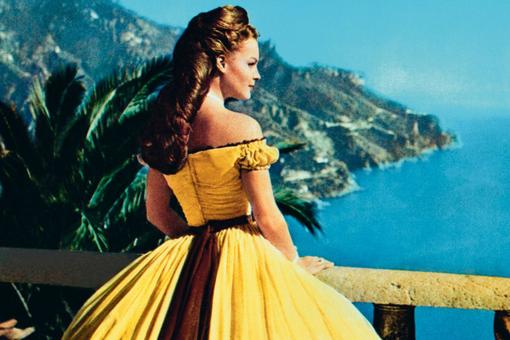 A photo of Romy Schneider as Empress Sisi in a sweeping yellow dress standing from a terrace looking out to sea.
