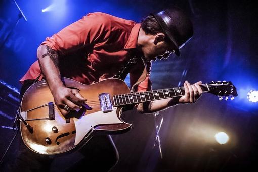 Photo of guitarist in red shirt and black hat playing guitar solo