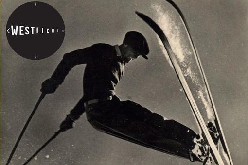 The historic black and white photo shows a skier jumping through the air, wearing a historic ski outfit and cap, with the WestLicht logo in the top left-hand corner. Scene for photography