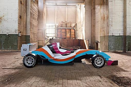 The photo shows an artistically designed racing car (Formula Renault 1.6) in the colors light blue, orange, white, pink and black. The car is located in a factory hall.