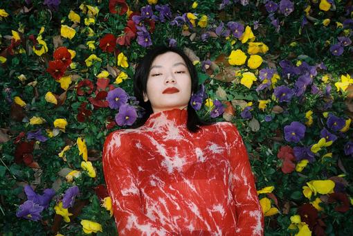 The picture shows a young Chinese woman in a red, white marbled top lying in a bed of purple, yellow and red pansies