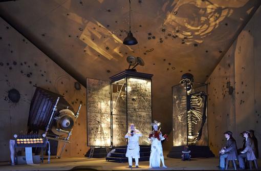 Imaginative scene with two main actors in white costumes in the foreground, a small group of spectators seated to the side, oversized glass display cases in the background, one with sheet music inside, one with an oversized skeleton