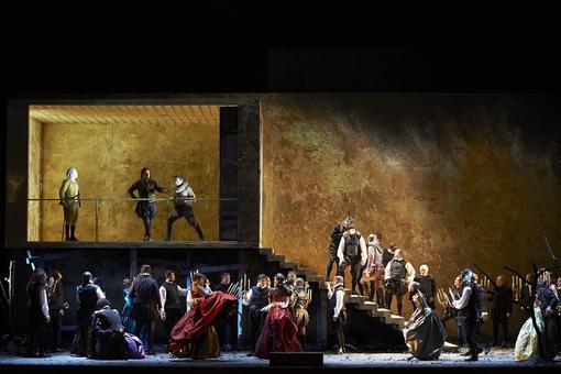 Scene photo from the opera Rigoletto: Rigoletto and the Duke of Mantua stand on a kind of balcony, below them the guests at a feast, men and women in historical costumes, holding candlesticks in their hands