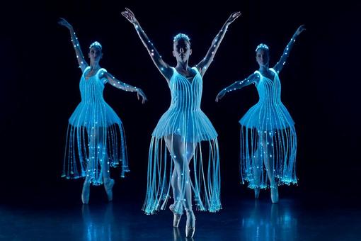 Three ballet dancers wearing costumes with fairy lights on a dark stage.