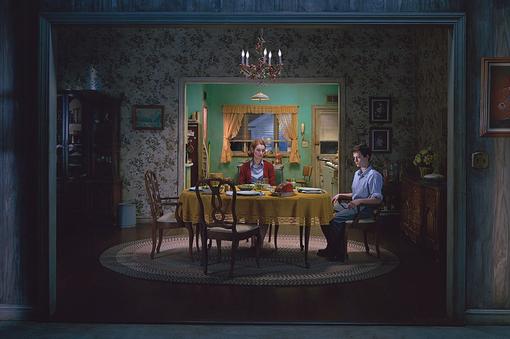 The photo shows a dining room with Biedermeier furniture. A woman and a man sit staring at a laid table, behind them you can see the kitchen in the style of the 1960s
